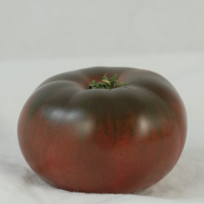 Almanac Planting Co Cherokee Purple Tomato. A heirloom, purple skinned tomato sitting on a white cloth with a white background. 