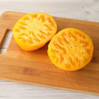 Almanac Planting Co Lemon Boy Hybrid Tomato. The tomato is sliced open into two pieces. The pieces are on top of a wooden cutting board.